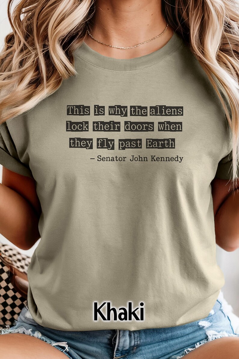 Op de afbeelding: Khaki-kleurig T-shirt met een citaat toegeschreven aan senator John Kennedy: "This is why the aliens lock their doors when they fly past Earth." Het citaat is in zwarte, typemachine-stijl lettertype. De kleur van het shirt is onderaan gelabeld met "Khaki".