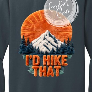 May include: Dark teal sweatshirt with a mountain scene. The design features an orange sun, a snow-capped mountain, and the text "I'D HIKE THAT" in orange, textured lettering. The sweatshirt is from Comfort Colors.