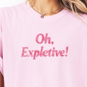 May include: A light pink t-shirt with the embroidered text "Oh, Expletive!" in a matching pink, fluffy font. The shirt has a crew neck and short sleeves. A silver necklace with a pendant is visible at the top.