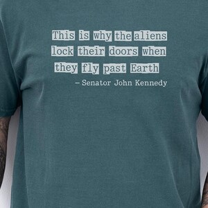 Op de afbeelding: Blauwgroen T-shirt met de quote "This is why the aliens lock their doors when they fly past Earth - Senator John Kennedy" in witte tekst.