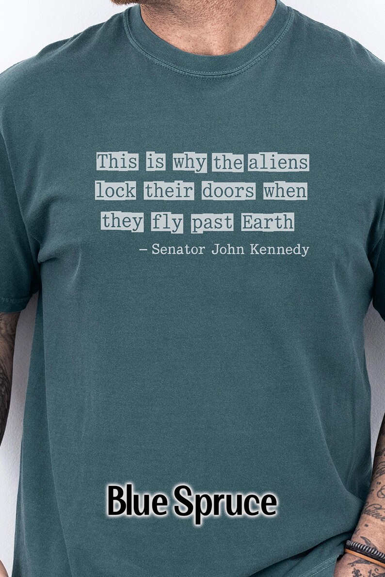 Op de afbeelding: Blauwgroen T-shirt met de quote "This is why the aliens lock their doors when they fly past Earth - Senator John Kennedy" in witte tekst.