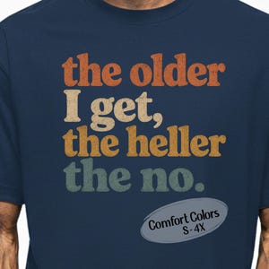 May include: Navy blue t-shirt with the text "the older I get, the heller the no." in orange, beige, yellow, and teal. The shirt includes a Comfort Colors label and size range S-4X.