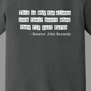Op de afbeelding: Grafietkleurig t-shirt met de quote "This is why the aliens lock their doors when they fly past Earth - Senator John Kennedy" in witte letters. Comfort Colors merk.