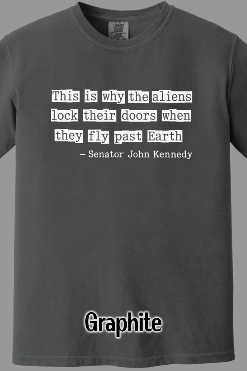 Op de afbeelding: Grafietkleurig t-shirt met de quote "This is why the aliens lock their doors when they fly past Earth - Senator John Kennedy" in witte letters. Comfort Colors merk.