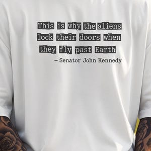 Op de afbeelding: Wit T-shirt met een citaat van Senator John Kennedy: "Dit is waarom de aliens hun deuren op slot doen als ze langs de aarde vliegen." Het woord "White" staat vetgedrukt onderaan het shirt.