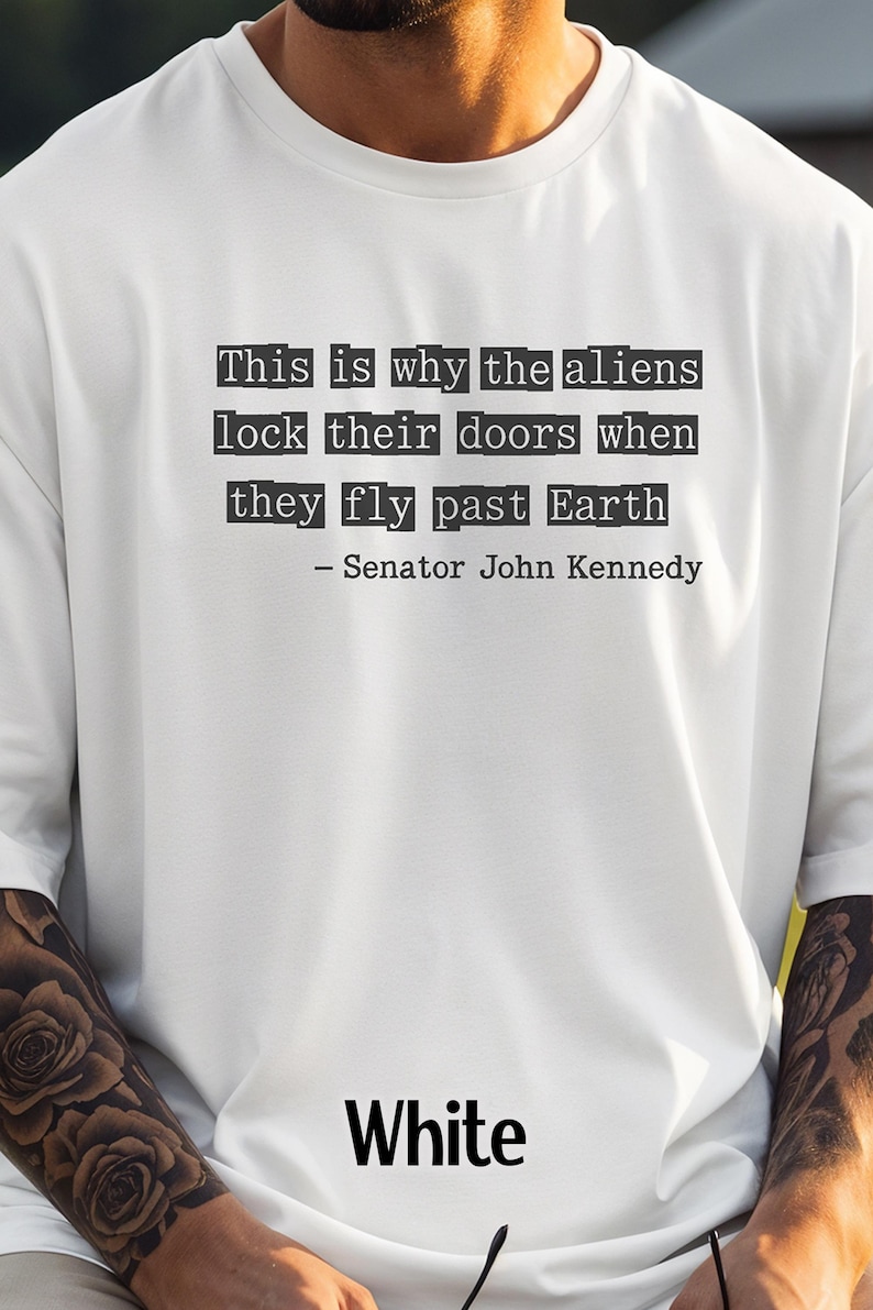 Op de afbeelding: Wit T-shirt met een citaat van Senator John Kennedy: "Dit is waarom de aliens hun deuren op slot doen als ze langs de aarde vliegen." Het woord "White" staat vetgedrukt onderaan het shirt.