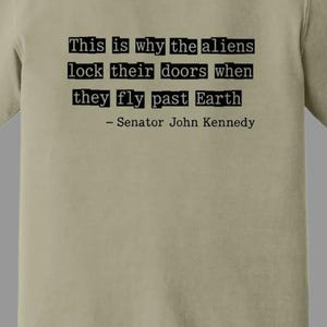 Op de afbeelding: Khaki T-shirt met de quote "This is why the aliens lock their doors when they fly past Earth - Senator John Kennedy" in het zwart. Gemaakt door Comfort Colors.