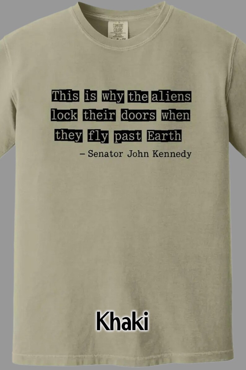 Op de afbeelding: Khaki T-shirt met de quote "This is why the aliens lock their doors when they fly past Earth - Senator John Kennedy" in het zwart. Gemaakt door Comfort Colors.