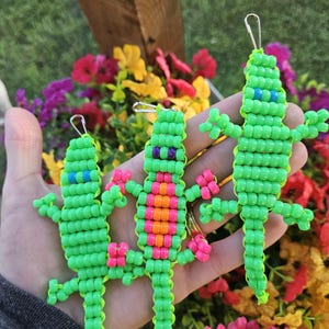 May include: Three lizard-shaped keychains crafted from neon green plastic pony beads. The left lizard has blue eyes, the center lizard has purple eyes and an orange and pink striped belly, and the right lizard has blue eyes. Each has a metal clasp.