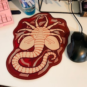 May include: A custom mousepad featuring a beige and brown graphic of a Xenomorph creature. The mousepad has a dark red border and is next to a black computer mouse.