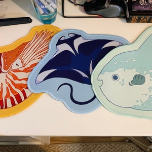 May include: Three decorative mouse pads with sea creature designs. One pad features a nautilus shell in orange and white on a yellow background. Another depicts a blue manta ray. The third pad shows a light blue sunfish.