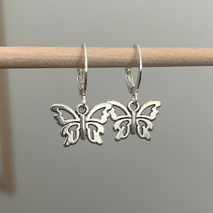 May include: A pair of silver butterfly earrings with a delicate, intricate design. The earrings are hanging from a wooden dowel.