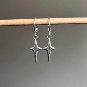 May include: A pair of silver-colored earrings. Each earring features a four-pointed star design with a hollow center. The earrings are suspended from a lever-back closure. The earrings are displayed on a wooden dowel against a neutral background.