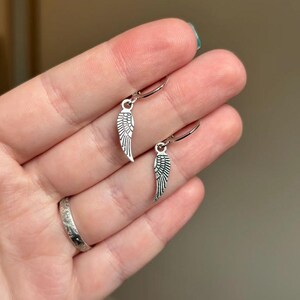 Pode incluir: Um par de brincos de argola de prata com um pequeno charme de asa de anjo de prata pendurado em cada argola.