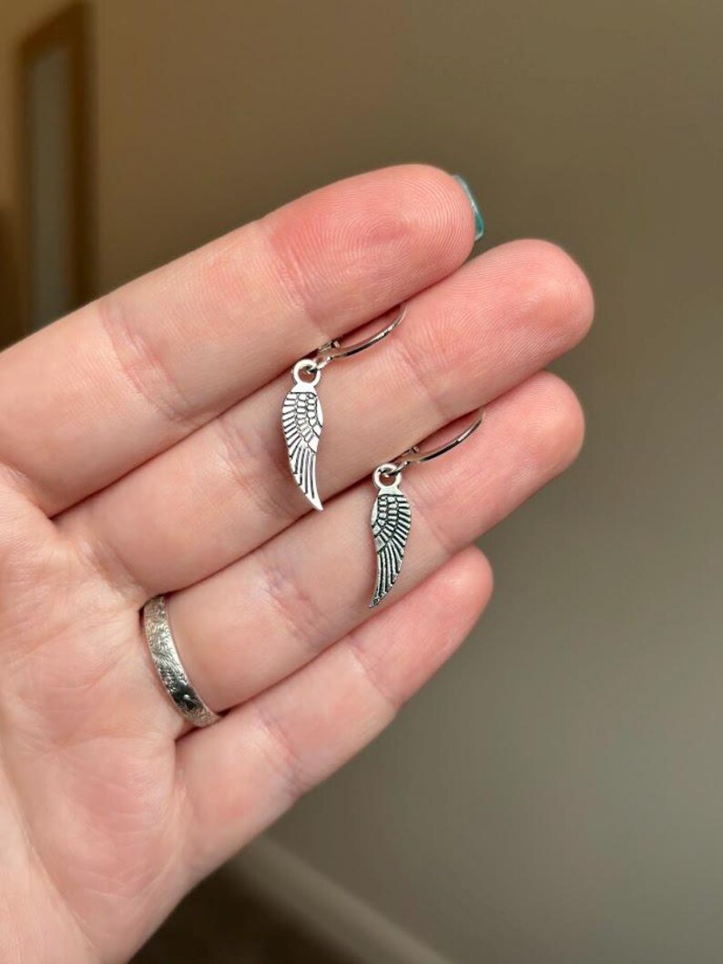 Pode incluir: Um par de brincos de argola de prata com um pequeno charme de asa de anjo de prata pendurado em cada argola.
