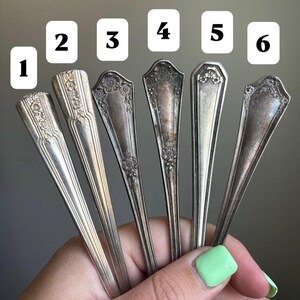 May include: Six antique silver spoons with ornate floral and geometric designs. Each spoon has a different pattern on the handle and bowl. The spoons are held in a hand with a light green painted fingernail.