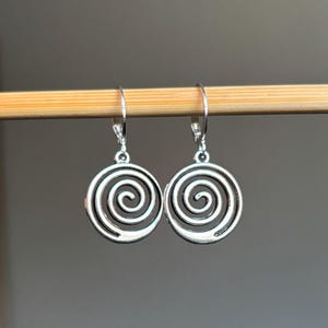 May include: Silver spiral earrings with a circular design. The earrings are hanging from a wooden surface.