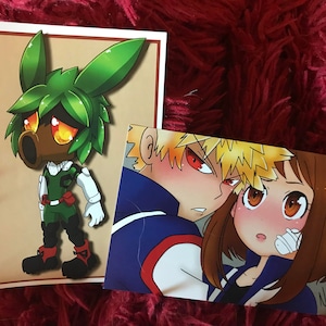 May include: Two anime-style illustrations printed on white cardstock. The first illustration features a green character with a brown face and large eyes. The second illustration features two characters, one with blonde hair and red eyes, and the other with brown hair and brown eyes. The text "Nomi Art" is in the bottom right corner of the second illustration.