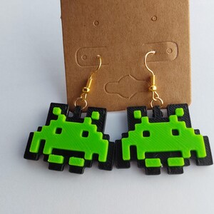 Könnte beinhalten: Ein Paar grüne und schwarze Space-Invader-Ohrringe. Die Ohrringe sind aus Kunststoff und haben ein pixeliges Design.