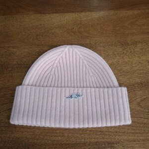 May include: A light pink ribbed knit beanie with a folded brim. A small embroidered design is on the front of the brim. The hat is made of a soft material and is suitable for cold weather.