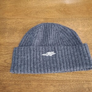 May include: A gray ribbed knit beanie with a folded brim. The beanie features a small, white embroidered armadillo logo on the front. The hat is made of a soft, warm material, perfect for cold weather.