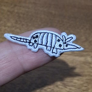 May include: A white embroidered patch in the shape of an armadillo. The armadillo has black details, including stripes, spots, and an outline. The patch is likely for clothing or accessories.