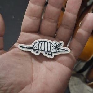 May include: A small, flat, armadillo-shaped patch or pin featuring a simple black line drawing on a light cream-colored background. The design depicts the armadillo in profile with a pointed snout, a segmented shell, and a long, thin tail.