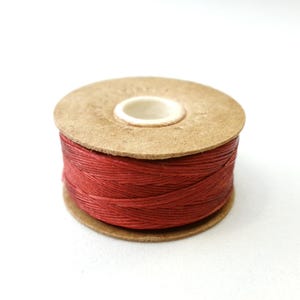 May include: A spool of red thread wound around a brown cardboard bobbin.