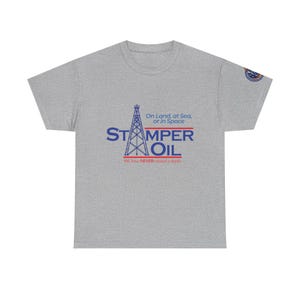 May include: A heather gray t-shirt with the text "STAMPER OIL" in blue, featuring an oil derrick graphic. The shirt also includes the phrase "On Land, at Sea, or in Space" and "We have NEVER missed a depth."