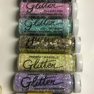 Op de afbeelding: Een set van acht kleine glazen flesjes gevuld met glitter. De flesjes zijn gelabeld met de kleuren van de glitter: zwart, roze, aqua, goud, geel, lavendel en wit. De flesjes zijn gelabeld met "Prisma Pastels Glitter" en "The Original Prisma Glitter".