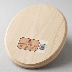 Könnte beinhalten: Eine helle, ovale Lindenholzplatte mit einer glatten, geschliffenen Oberfläche. Auf der Vorderseite befindet sich ein rechteckiges Produktetikett mit dem Text Walnut Hollow, Basswood Plaque, 8 x 10 Oval sowie einem Barcode und Details.
