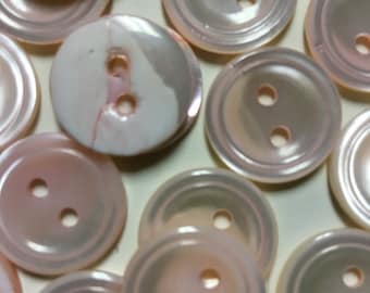 May include: A collection of two-hole pearl buttons in various shades of pink and white. The buttons have a smooth, shiny finish and are arranged in a random pattern.