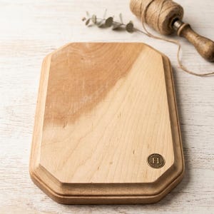 Könnte beinhalten: Eine rechteckige Holzplatte mit abgeschrägten Kanten und einer hellbraunen, natürlichen Holzmaserung. In der unteren rechten Ecke ist ein kleines, messingfarbenes Metallemblem eingelassen, das den Buchstaben H in einem Rand zeigt.