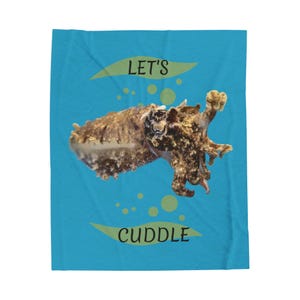 Puede incluir: Una manta azul con una ilustración de una sepia. La manta tiene las palabras "LET'S" y "CUDDLE" en una fuente lúdica, con acentos amarillos. La sepia es marrón y beige.