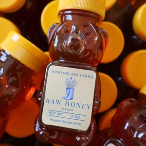 May include: Several bear-shaped bottles of raw honey with yellow lids. Each bottle has a label that reads "Bowling Bee Farms Raw Honey" and "Net Wt. 8 oz."