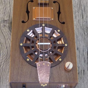 May include: A cigar box guitar with a copper sound hole and a copper pickguard. The guitar has a wooden body and a wooden neck. The guitar has a volume knob and a jack for an audio cable.