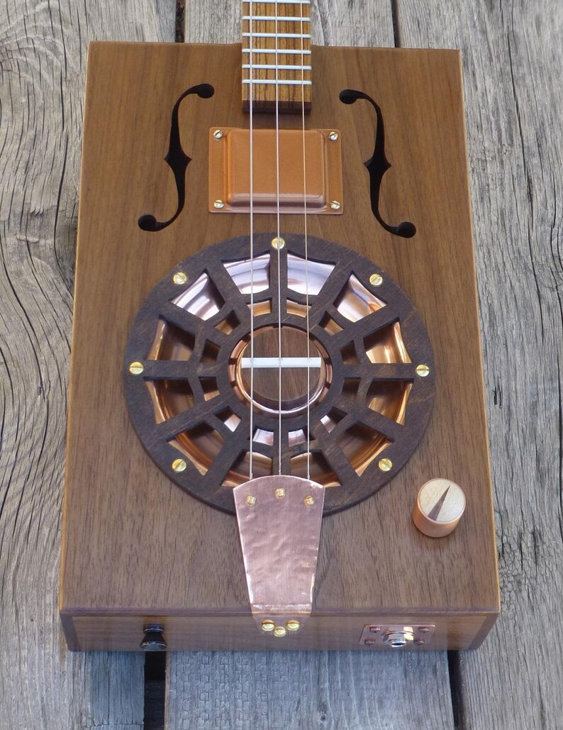 May include: A cigar box guitar with a copper sound hole and a copper pickguard. The guitar has a wooden body and a wooden neck. The guitar has a volume knob and a jack for an audio cable.