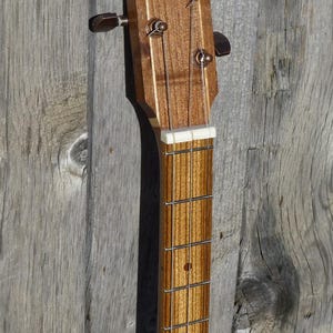 May include: Close-up of the headstock of a wooden guitar with a light brown finish. The headstock has four tuning pegs with a white plastic nut and a white plastic fret.