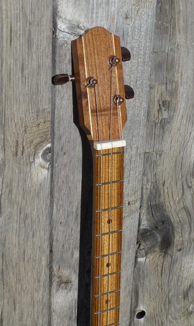 May include: Close-up of the headstock of a wooden guitar with a light brown finish. The headstock has four tuning pegs with a white plastic nut and a white plastic fret.