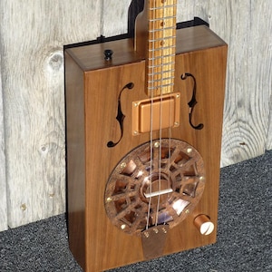 May include: A wooden cigar box guitar with a copper resonator and a wooden neck. The guitar has a single volume knob and a black switch.