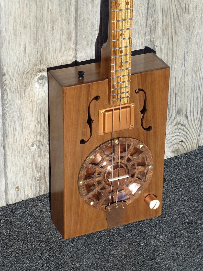 May include: A wooden cigar box guitar with a copper resonator and a wooden neck. The guitar has a single volume knob and a black switch.
