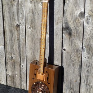 May include: A cigar box guitar with a wooden body and a copper resonator. The guitar has a wooden neck and a set of tuning pegs at the top of the neck.