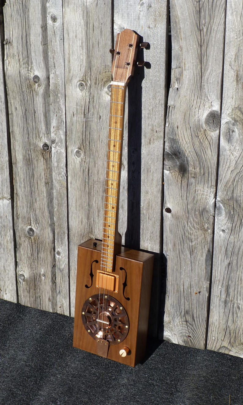 May include: A cigar box guitar with a wooden body and a copper resonator. The guitar has a wooden neck and a set of tuning pegs at the top of the neck.