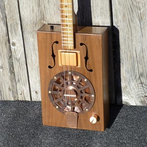 May include: A wooden cigar box guitar with a copper resonator and a walnut body. The guitar has a single pickup and a volume knob.