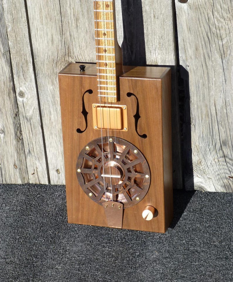 May include: A wooden cigar box guitar with a copper resonator and a walnut body. The guitar has a single pickup and a volume knob.