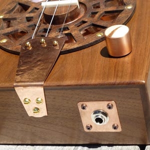 May include: A wooden cigar box guitar with copper accents, a round sound hole with a decorative metal grille, and a copper output jack.