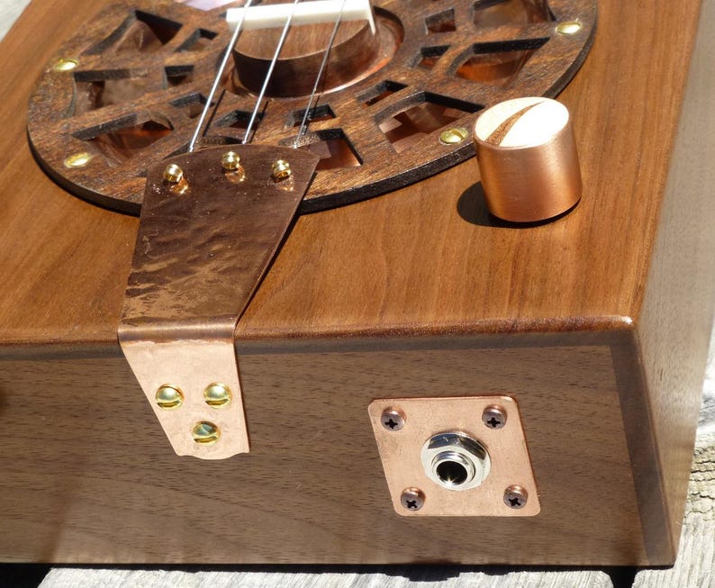 May include: A wooden cigar box guitar with copper accents, a round sound hole with a decorative metal grille, and a copper output jack.