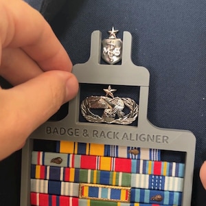 May include: A gray plastic badge and rack aligner tool holding military ribbons and insignia. The tool has the text "BADGE & RACK ALIGNER" printed on it. The ribbons are in various colors, including red, blue, yellow, and green.