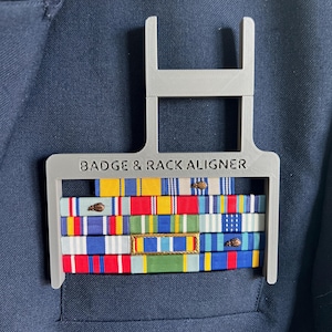 May include: A gray plastic badge and rack aligner holding several military ribbons with various colors. The text "BADGE & RACK ALIGNER" is printed on the aligner. The ribbons are attached to a navy blue jacket.