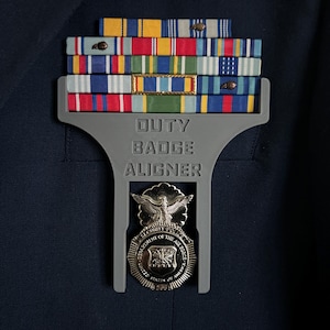 May include: A gray Duty Badge Aligner holding several military ribbons and a Department of the Air Force Security Police badge. The ribbons are in various colors, including red, blue, yellow, and green. The badge features an eagle and the text "United States Air Force".